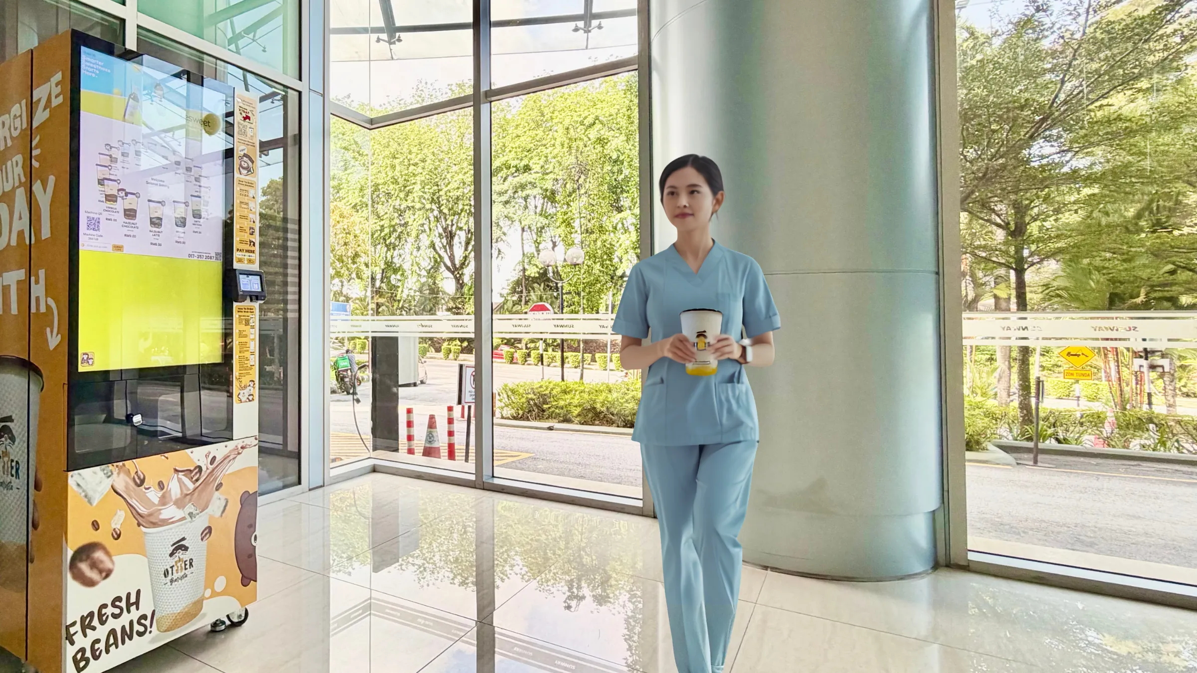A nurse in scrubs walking through a hospital corridor with a takeaway coffee, past an Otter Barista machine